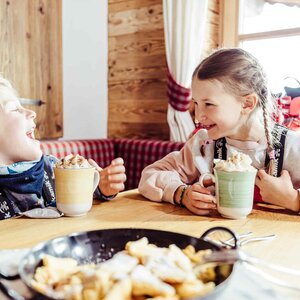 Zwei Kinder sitzen in der Skihütte, trinken heiße Schokolade mit Sahne und haben vor sich eine Pfanne voller Kaiserschmarrn stehen | © Alpbachtal Tourismus | shootandstyle 